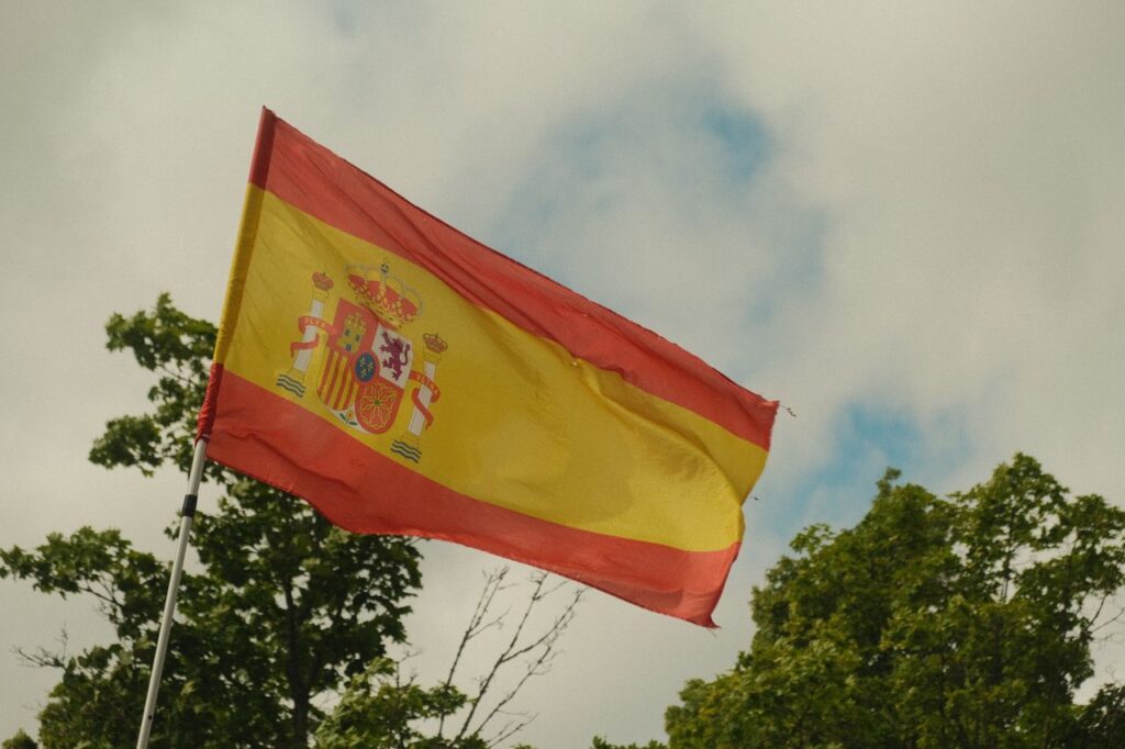 Spanish flag fluttering against a backdrop of trees and cloudy sky.